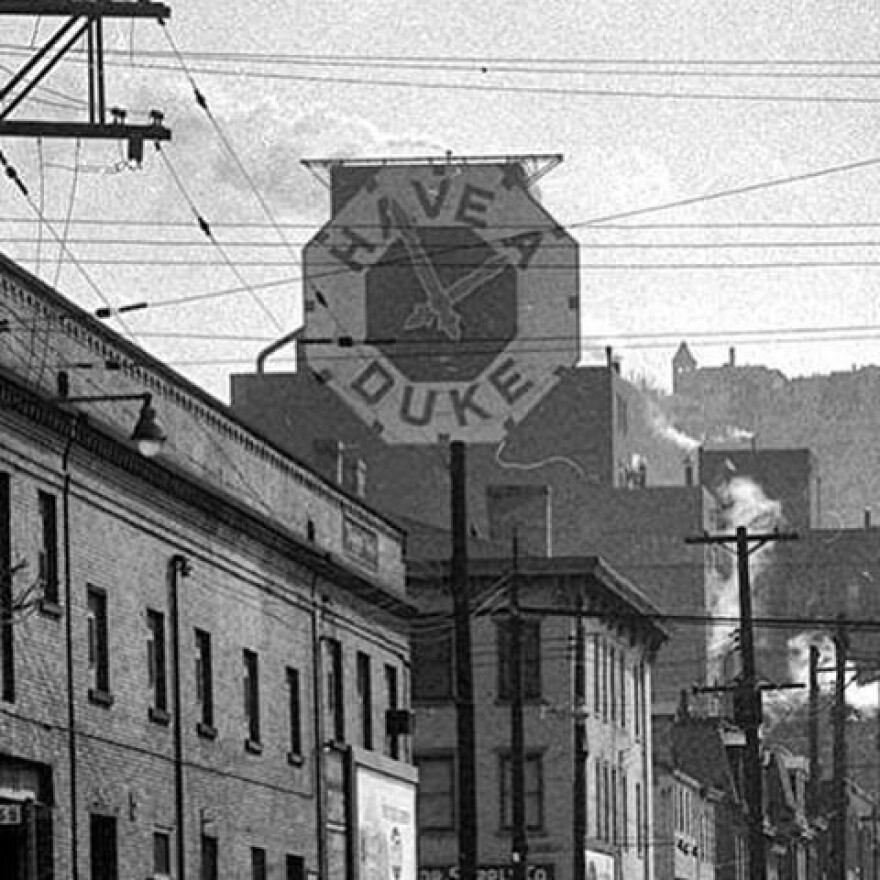 An old black-and-white photo of a massive clock on the side of a building with text reading "Have a Duke."
